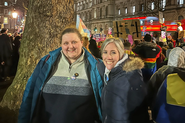 Foreground: LGBT+ Lib Dems Chair, Charley Hasted stood beside Daisy Cooper, MP for St Albans, Deputy Leader of the Liberal Democrats and Spokesperson for Health and Social Care.  Background: Crowd of demonstrators holding placards and trans flags