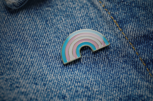 Enamel trans pin badge on a light denim jacket. Image brightness has been darkened.